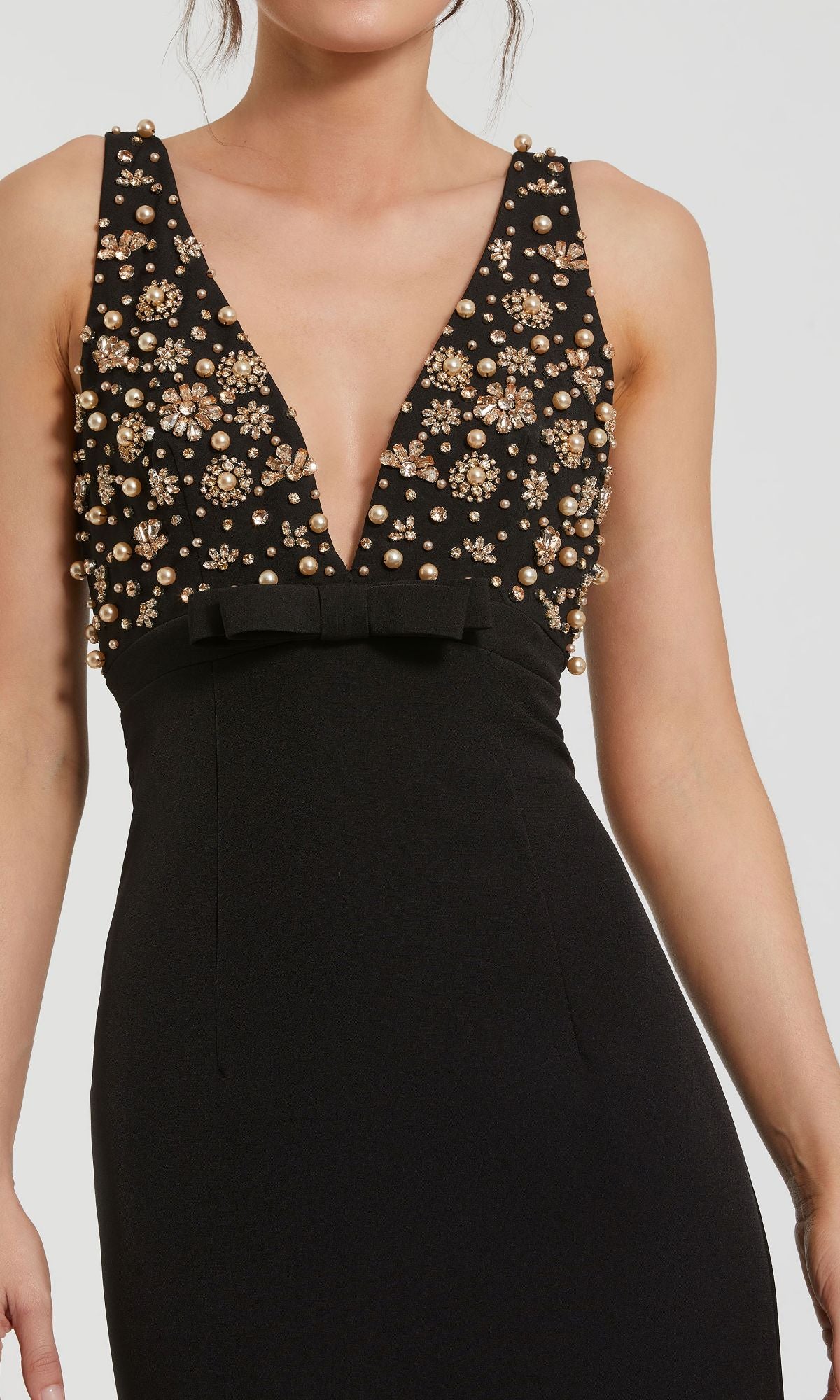 A woman wears the Mac Duggal 2278: a sleeveless black beaded-bodice long prom dress with a deep V neckline, empire waist, pearl and rhinestone floral details, and a delicate black bow.